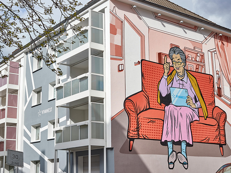 Ein modernisiertes Gebäude. An eine Hauswand wurde ein großes Bild gemalt. Darauf zu sehen ist eine ältere Dame, die in ihrem Wohnzimmer auf einem roten Sofa sitzt und ein Tablet mit VIVAWEST-Schriftzug in der Hand hält