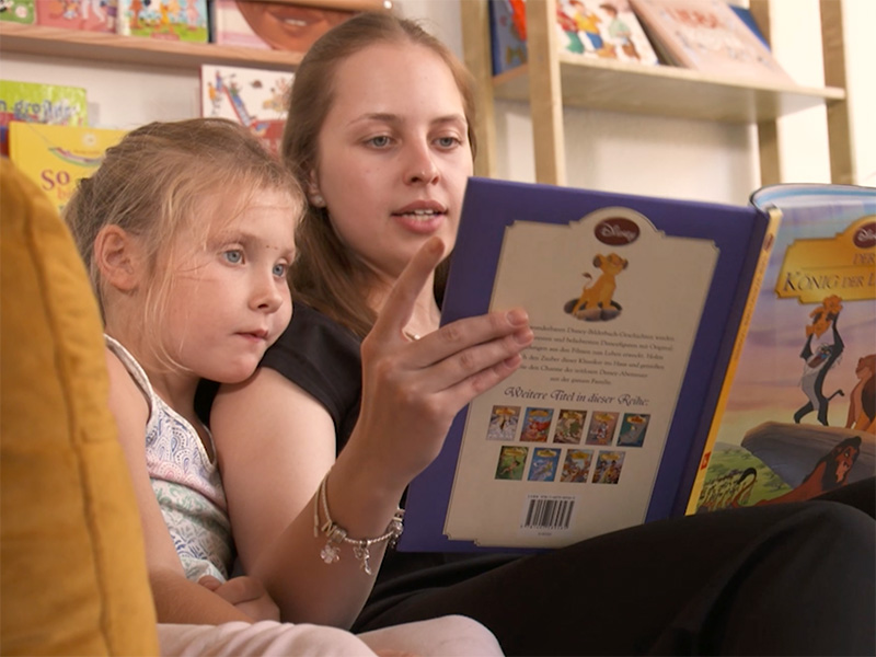 Eine Frau und ein Mädchen sitzen auf einem gelben Sofa. Das Mädchen hat seinen Kopf an die Schulter der Frau gelehnt. Die Frau liest dem Mädchen aus dem Buch 'König der Löwen' vor.