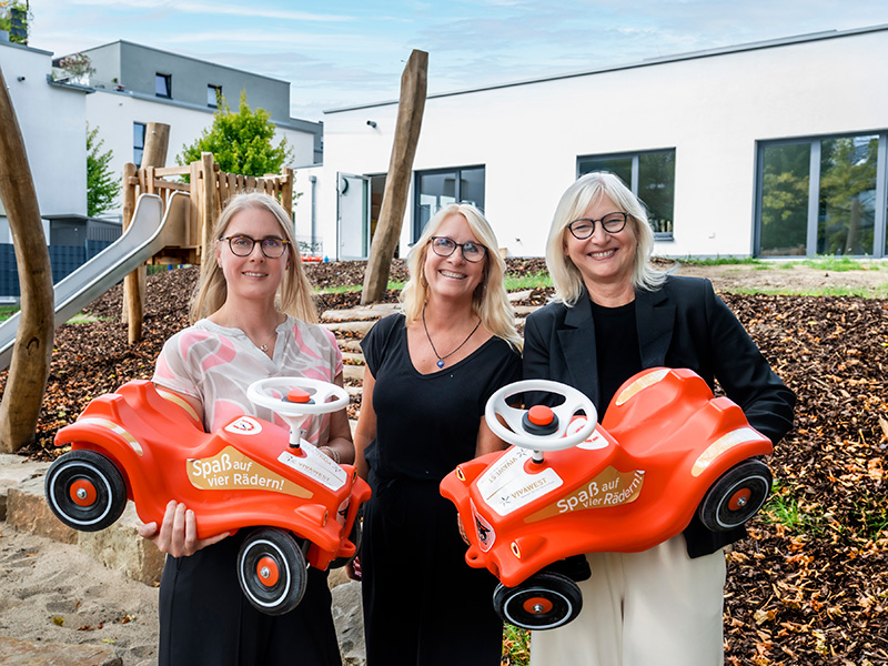 Drei Frauen stehen vor einer neu eröffneten Kita, im Hintergrund ist ein Spielplatz mit Rutsche zu sehen. Sie lächeln in die Kamera. Zwei der Frauen halten jeweils ein rotes Bobbycar hoch.