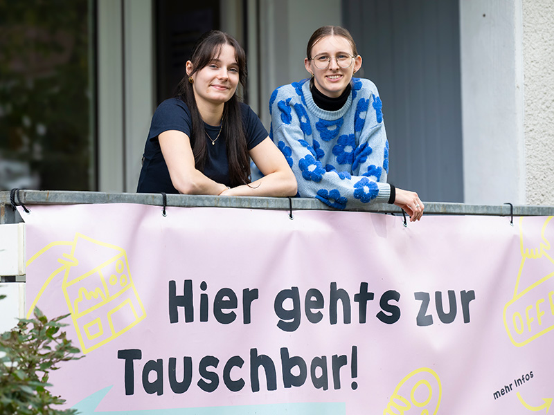Zwei junge Frauen stehen auf einem Balkon und lächeln in die Kamera. Am Balkongeländer hängt ein Banner auf dem 'Hier geht es zur Tauschbar!' steht.